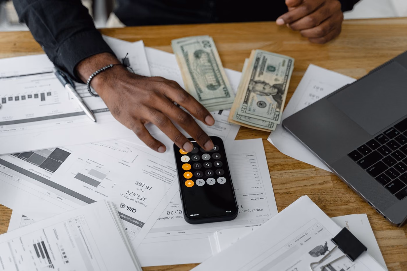 Mão calculando orçamento com calculadora, notas de dinheiro e laptop ao fundo